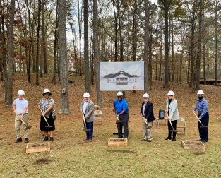 Groundbreaking ceremony at Camp Fletcher for the new dining hall. Seven people in hard hats stand in a row outdoors, each holding a shovel. Behind them is a poster rendering of the planned building, set against a wooded backdrop